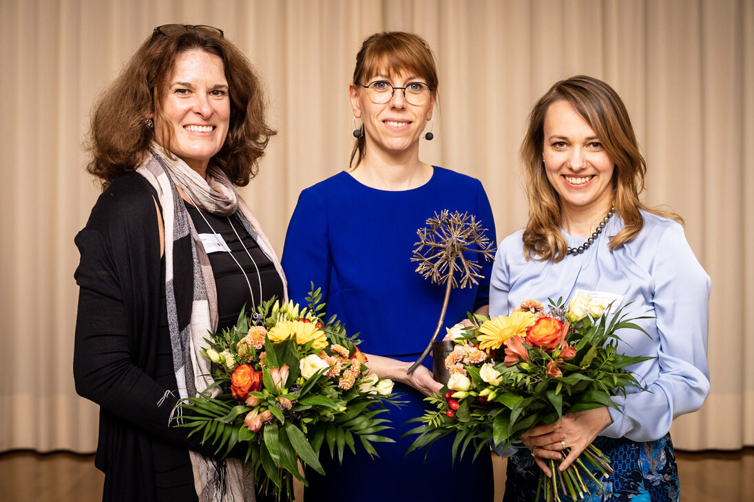 Drei Frauen stehen nebeneinander und lächeln in die Kamera, Frauen links und rechts halten jeweils einen großen Blumenstrauß