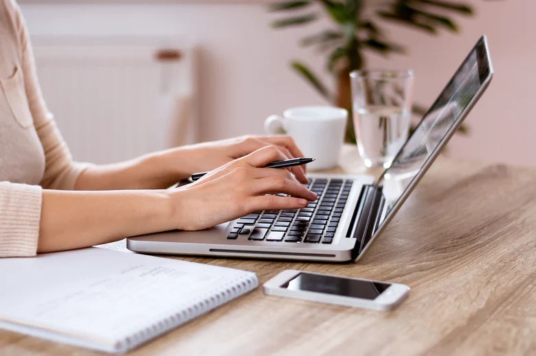 Nahaufnahme von Händen, die an einem aufgeklappten Laptop an einem Holztisch tippen, daneben Notizblock mit Stift, Smartphone, Glas Wasser und Kaffeetasse in heller, büroähnlicher Umgebung.