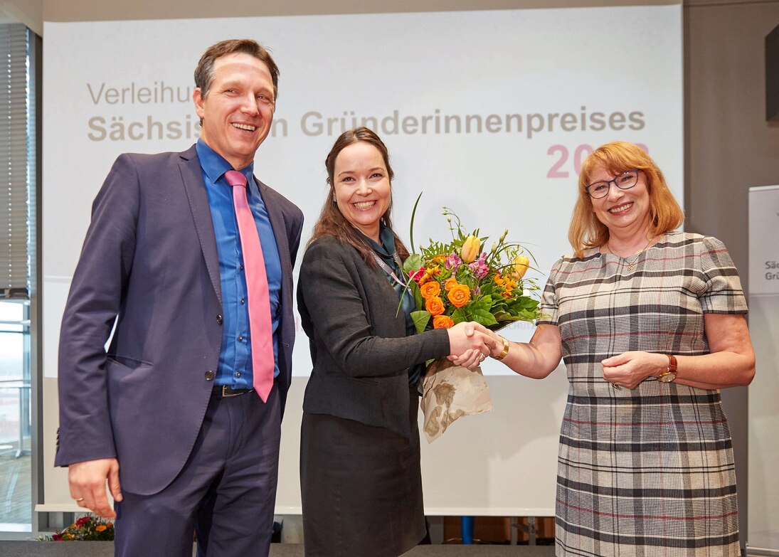 Ein Mann und zwei Frauen stehen nebeneinander und lächeln, der jungen Frau in der Mitte werden bunte Blumen überreicht