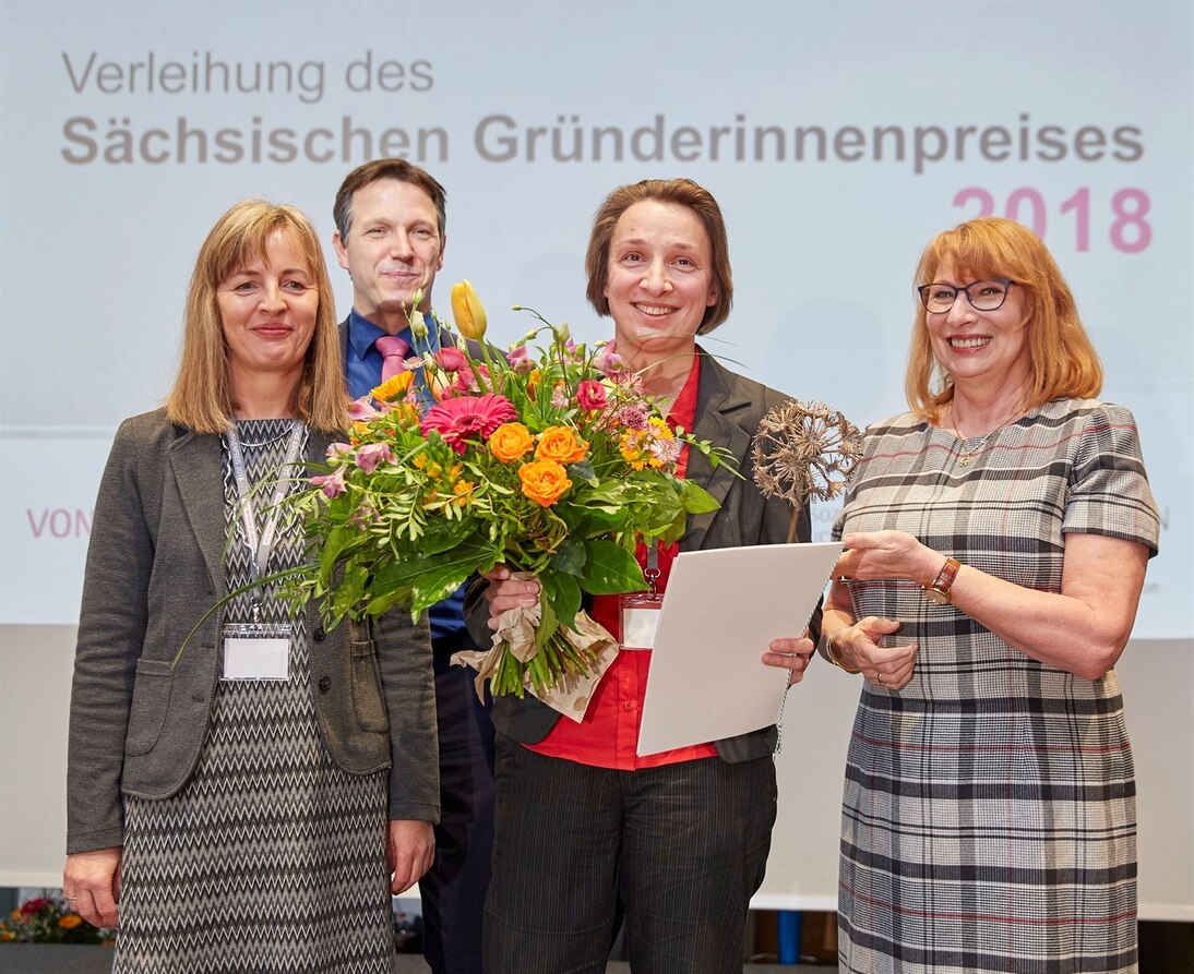 Drei Frauen stehen nebeneinander und lächeln, hinter ihnen steht ein Mann, die Frau in der Mitte hält einen großen Blumenstrauß, Urkunde und eine Figur in Form einer Pusteblume